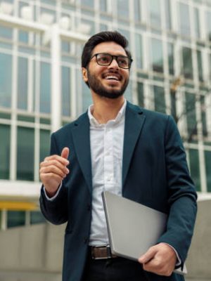 istockphoto-1448167415-612×612 Smiling businessman in suit with laptop near modern office building is looking at side